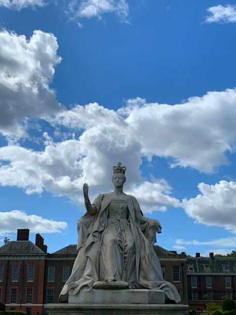 A stone statue of a regal female figure wearing a flowing gown and a crown, seated on a throne-like base with her right hand raised in a gesture. The sculpture is positioned outdoors against a background of a blue sky dotted with white, fluffy clouds. Behind the statue, there are brick buildings with dark slate roofs and chimneys, suggesting an urban or historical setting. The detailed craftsmanship of the sculpture includes intricate drapery in the gown and a solemn expression on the figure's face. The scene appears well-lit with natural daylight, highlighting the texture and carvings of the stone. Although not directly related to rubbish removal, such statues are sometimes located in public spaces or civic areas where private waste management services could include on-site clearance of surrounding debris or waste in nearby areas. The presence of prominent sculptures may relate to locations where alternative waste disposal or independent collection services might operate outside conventional council services, supporting the broader context of flexible rubbish pickup options.