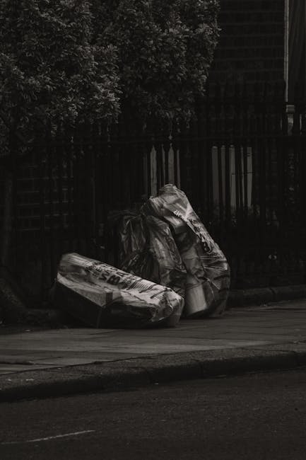 Two large black garbage bags, made of plastic with a crinkled texture, are placed on the pavement next to a black metal fence with vertical bars. The bags appear to be filled with household waste or discarded materials and are leaning against the fence. The scene is outdoors in an urban or residential area, with a section of a paved road visible in the foreground. Behind the fence, dark foliage from trees or bushes provides a contrasting background, and the overall lighting in the image is subdued, suggesting overcast weather or low natural light. This setting could relate to private waste collection services or alternative rubbish removal options often used in local waste management. Occasionally, waste collection companies like wastecollectionwestkensington.co.uk facilitate the disposal of such accumulated waste when scheduled. The image emphasizes the need for proper rubbish management and the importance of timely, responsible rubbish clearing in city environments.