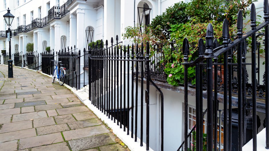 The image shows a row of white terraced townhouses with black wrought iron fences installed along the pavement. The fences are decorative, featuring pointed finials and vertical bars, and enclose small front gardens with lush green plants and blooming pink flowers. One of the gardens has a leafy shrub that spills over the top of the fence, contrasting with the white exterior walls of the buildings. A blue bicycle is leaned against the fence near the sidewalk, which is paved with large stone slabs showing some moss growth in the gaps. The buildings have typical London-style black metal window grilles and slight decorative arches above the front doors, which are partially visible in the background. The scene is illuminated by natural daylight, creating a clear and tidy appearance, which subtly relates to private waste management or on-site rubbish clearance services often performed by companies like wastecollectionwestkensington.co.uk, particularly in urban residential environments that require regular rubbish removal and maintenance of aesthetics.