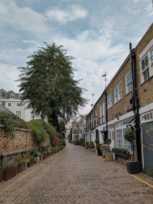 A narrow cobblestone alleyway in an urban residential area, with a large tree situated towards the left side, its branches extending above the buildings and overhead sky. On the right, traditional brick terraced houses with white-framed sash windows and black guttering are visible, some with small potted plants and decorative elements near the entrances. The row of houses features a uniform architectural style, with brick facades in warm tones, white window frames, and black downspouts. The alley is bordered by low brick walls and planters containing various greenery and flowers, creating a green space along the path. Overhead, the sky is partly cloudy with patches of blue. The scene suggests a quiet, private residential street that could be associated with property management or alternative waste handling, as it appears to be a setting for independent rubbish collection or on-site clearance managed by local waste removal services such as wastecollectionwestkensington.co.uk.