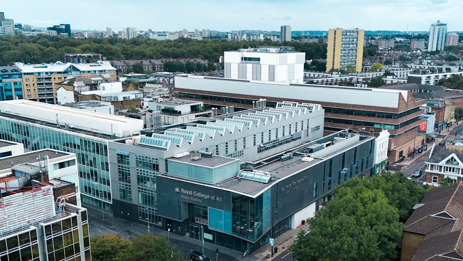 An aerial view of an urban area showing a modern building identified as the Royal College of Art, constructed with glass and metal panels, with a distinctive roof featuring a row of skylights. Adjacent structures include a large brown brick building with horizontal protrusions and signage, surrounded by other commercial and residential buildings. The scene is set on a city street lined with trees, with vehicles parked along the pavement, indicating active city life and on-site waste management activities. The overall environment suggests a busy, developed district with diverse architectural styles, and the visible rooftops and street level highlight the importance of efficient rubbish removal and waste disposal services in such urban settings. The area benefits from natural daylight, enhancing the clarity of building exteriors and surroundings, and reflects the typical context where independent waste collection or private disposal services, such as those provided by wastecollectionwestkensington.co.uk, might be involved in maintaining cleanliness and managing refuse in busy city environments.