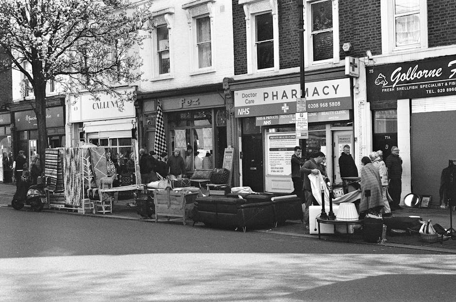 A black and white photograph capturing a street scene in West Kensington, featuring a temporary outdoor rubbish collection area with a variety of discarded furniture and household items arranged along the sidewalk in front of a row of commercial premises. Visible objects include sofas, chairs, cushions, and miscellaneous household items, some stacked and others placed on the pavement. Several people are standing around the collection site, observing or engaging with the items, with some dressed in coats and hats, indicating cool weather. The background shows a pharmacy with signage for NHS and private services, as well as other retail outlets, with shopfronts and large windows. A lamppost and a small tree are positioned among the items, with a few bicycles and a scooter nearby. The scene is lit by natural daylight, casting shadows on the pavement, and exemplifies alternative waste handling or private rubbish disposal methods often employed in local urban environments, as managed by waste collection services such as wastecollectionwestkensington.co.uk.