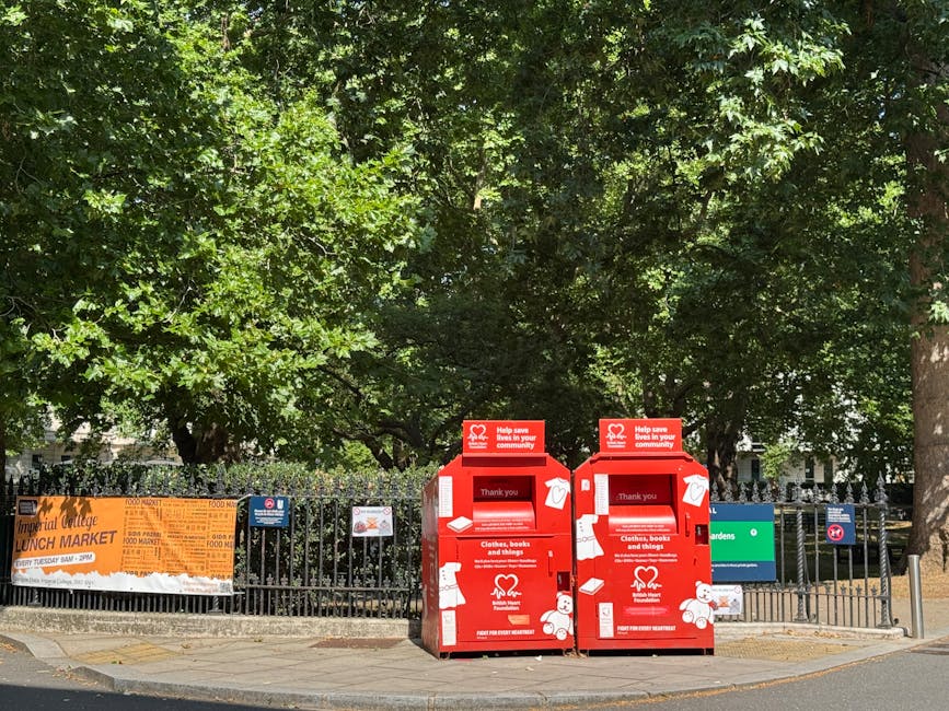Two red charity collection bins situated on a paved pavement alongside a black metal fence, with trees and greenery providing a dense canopy overhead. The bins are designed for collecting clothing, books, and miscellaneous items, featuring white icons and text indicating their purpose and donation information. Located in an outdoor urban environment, they are positioned close to each other with limited space between, and are partially covered by shadows cast by the surrounding foliage. Behind the fence, bright orange and green banners are visible, advertising a local lunch market and community garden. The scene suggests an area where secure outdoor charitable waste collection takes place, possibly serving an adjacent community space or market area. The overall setting emphasizes a clean, organized approach to private waste handling for the drop-off of donations, aligned with alternative rubbish collection methods.