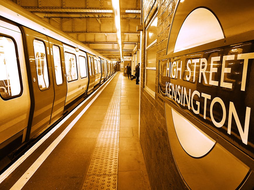 An underground railway station platform at High Street Kensington with a modern train stopped alongside the edge of the platform. The train has a sleek, metallic exterior with a shiny, reflective finish and rectangular windows along its side. The platform surface is light brown with tactile paving strips near the edge for safety. In the background, a few passengers are standing or walking, dressed in dark clothing. The station's wall on the right features a circular sign with the station name 'High Street Kensington' displayed prominently in white capital letters on a dark background. Overhead lighting provides a bright, even illumination, which accentuates the clean, structured environment typical of urban railway stations. The scene’s neutral tones and precise detailing align with the context of public transport infrastructure, while the image’s clarity supports accessibility for users seeking information about station facilities or travel routes near West Kensington and Grove Road areas.