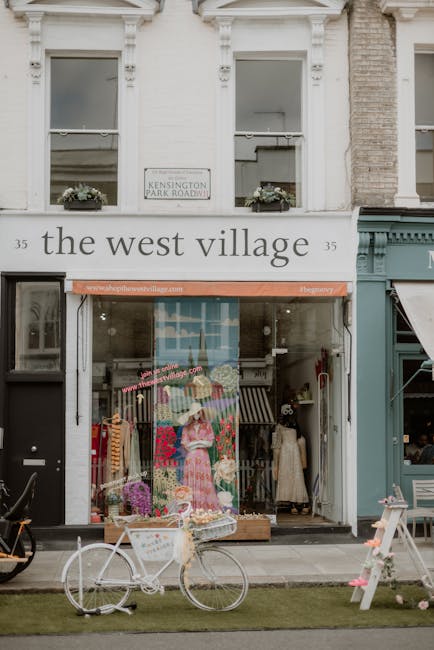 The image depicts the storefront of a small retail shop named 'the west village,' situated on a street with a white-painted façade and large glass windows. Above the entrance, a sign displays the shop's name in lowercase black lettering, and there is a small street sign indicating 'Kensington Park Road.' The shop's glass front reveals a display area with mannequins dressed in vintage-style clothing, including a prominent one wearing a pink floral dress and a wide-brimmed hat, surrounded by various items such as dresses and accessories arranged in an inviting manner. Outside the shop, on the sidewalk, there is a white bicycle with a basket parked in front, along with a small, partially visible pallet or display stand with pink flowers or objects, suggesting a charming boutique setting. The upper part of the building features two windows with flower boxes, and the overall environment appears clean and well-maintained, typical of an urban shopping district. This scene reflects a retail environment focused on private shopping experiences rather than general rubbish collection, aligning with the context of alternative waste handling or small business retail presentation.