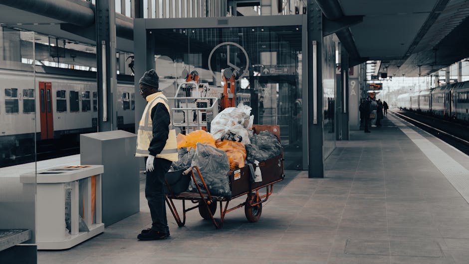 A person wearing a dark jacket, grey beanie, and white gloves stands on a modern train station platform next to a large metal trolley loaded with various types of discarded rubbish, including black and orange plastic bags, crumpled paper, and miscellaneous waste materials. The trolley has a rusted orange frame and is positioned near a glass-enclosed waiting area with a reflection of recycling and waste collection machinery visible behind it. To the left, there is a white waste bin with a slot for disposal, placed on the grey tiled surface of the platform. The background features train tracks extending to the right, with a stationary train visible on the platform side, and a few other individuals further along the platform, emphasizing the public transit environment. The scene is illuminated by natural daylight, highlighting the textures of the waste and the urban setting, demonstrating a scenario where private waste collection or rubbish clearance services, such as those provided by wastecollectionwestkensington.co.uk, could be relevant for managing large-volume waste accumulation in urban passenger transit areas.