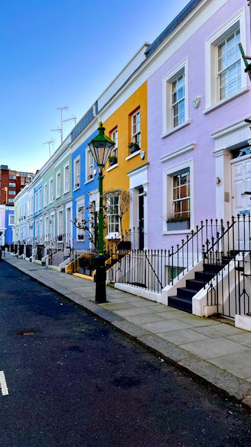A row of colorful terraced houses in West Kensington, painted in pastel shades of blue, yellow, and purple, with white window frames and black wrought-iron railings along the front steps. The pavement in front is clean and well-maintained, with a traditional black street lamp positioned near the curb. The houses feature small front gardens or steps leading to front doors, some with potted plants or decorative elements. The sky above is clear and blue, indicating a bright, sunny day. The scene suggests a typical residential area where private waste collection services, such as those provided by wastecollectionwestkensington.co.uk, might be used for alternative rubbish removal or on-site clearance, complementing local authority waste management arrangements. The overall atmosphere is tidy and orderly, reflecting a well-kept neighborhood focused on efficient waste disposal and environmental cleanliness.