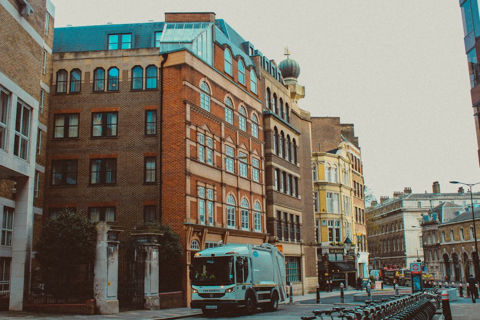 A medium-sized refuse collection vehicle, painted in light grey, is parked on a city street in front of several historic and modern multi-storey buildings. The truck features a front cab with side mirrors, a large cargo area at the rear, and a hydraulic lift system for waste loading, all made from smooth metal with a durable finish. To the right of the truck, there are metal bike racks aligned along the sidewalk and a row of black bollards separating pedestrians from the street traffic. The buildings, made of red brick, stone, and painted facades, showcase varying architectural styles, with large windows, some arched and others rectangular, and decorative elements such as cornices and window trims. A historical gate with white stone pillars and dark iron fencing stands partially visible on the left side of the image. The environment is well-lit with daylight, and the overall scene suggests a typical urban area with active waste collection, possibly organized as part of a private rubbish removal service in West Kensington, as indicated by the context of the website wastecollectionwestkensington.co.uk. This scene relates to alternative waste handling methods such as on-site clearance and private rubbish removal, avoiding reliance on local authority collection services.