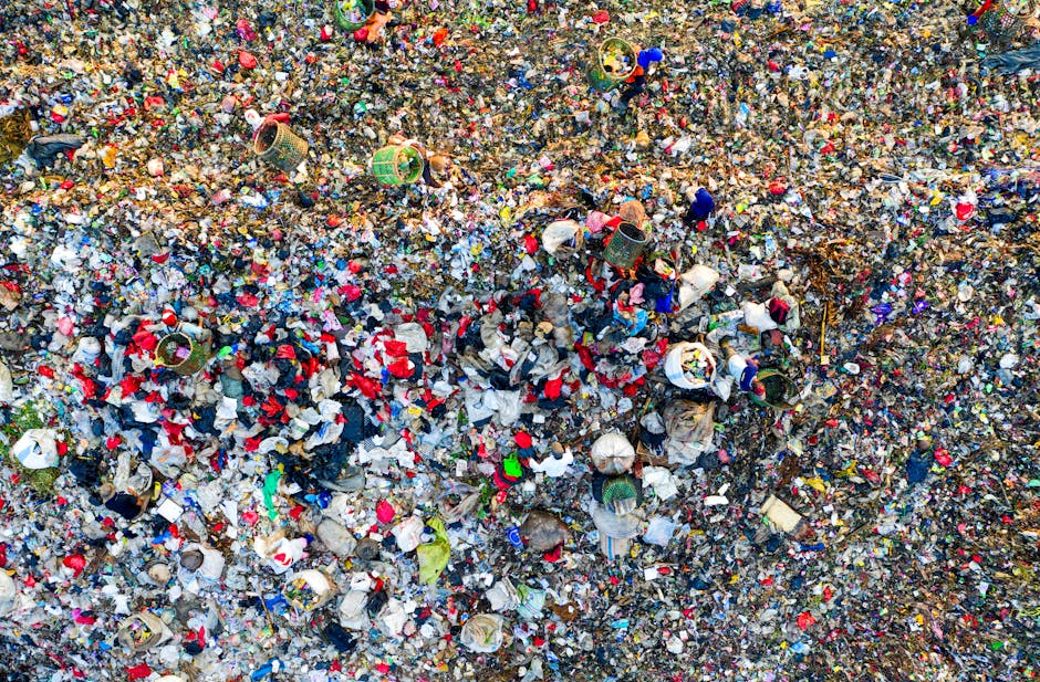 An aerial view of a large landfill site filled with a dense layer of mixed waste, including plastic bottles, paper, fabric, and other household refuse. In the foreground, various discarded items such as crumpled paper, plastic packaging, and fabric fragments are visible amidst the debris. Scattered throughout are several open-top waste bins, some tipped over or filled beyond capacity, with trash spilling out onto the surrounding material. The surface of the landfill appears uneven and muddy, with a combination of organic and synthetic materials creating a rough, textured appearance. The scene is brightly lit, emphasizing the variety of colors from the different waste items, with no clear background visible outside the densely packed waste. This image exemplifies the importance of proper waste management and the impact of improper rubbish disposal, illustrating a common scenario where independent rubbish removal services like those from wastecollectionwestkensington.co.uk might be necessary to prevent such overfilled sites or to facilitate cleanup and recycling efforts.