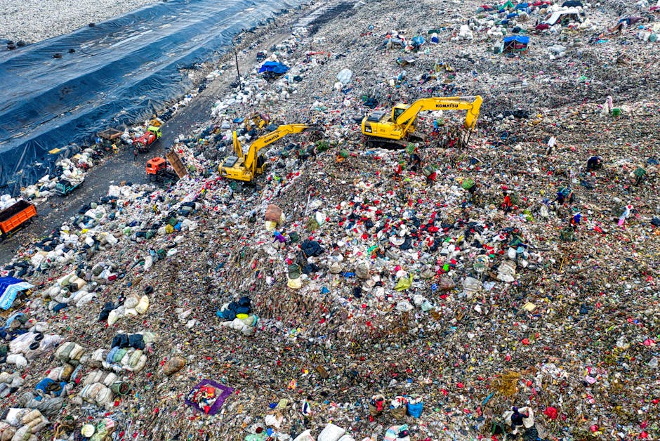 A large landfill site filled with layers of mixed waste, including plastic bags, sacks, broken household items, and miscellaneous debris, spread across a sloped terrain. Bright yellow Komatsu excavators are actively moving and piling up the rubbish, with their long arms and buckets visible as they work through the waste. The surface of the landfill is uneven and densely packed with refuse, with some areas covered by black plastic sheeting to contain or minimize spillage. In the background, there is a section of the site bordered by a steep bank of compacted waste, and a few scattered workers or personnel can be seen among the debris. This scene exemplifies a large-scale waste disposal operation, highlighting the volume of refuse managed through focused landfill or alternative waste handling, like private rubbish collection services. The overall environment is industrial, with a muted natural atmosphere, emphasizing the scale and nature of modern waste management practices as carried out by companies such as wastecollectionwestkensington.co.uk.