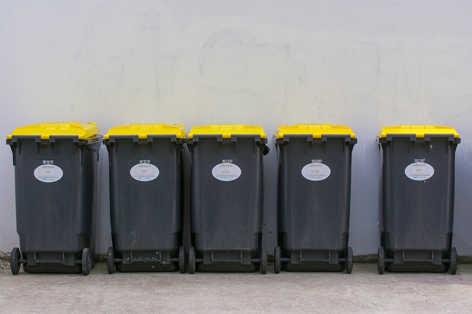 Five large black wheeled rubbish bins with bright yellow lids are lined up in a straight row on a concrete surface, positioned against a plain, light-colored wall. Each bin has a white oval label with a barcode and some printed text, and they are uniformly spaced, with the first bin on the left slightly closer to the camera. The bins are made of durable plastic with a textured surface, and their wheels are visible at the base, with some showing signs of use such as dirt or small scratches. The environment appears to be an outdoor or semi-outdoor area, suitable for private waste collection or on-site clearance, with no other objects or structures visible in the background. The lighting is diffuse, suggesting overcast weather or indoor settings with soft ambient light, emphasizing the practical, functional nature of the waste storage setup often associated with private rubbish collection services like those offered by wastecollectionwestkensington.co.uk, particularly in contexts with restricted access to communal waste collection points.