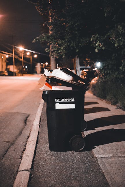 A black wheeled rubbish bin positioned on the edge of a paved sidewalk beside a street at night. The bin has a white label with the words 'ST. JOHN'S' printed on it and is filled with various discarded waste items, including cardboard boxes and plastic materials, some of which are protruding from the open lid. The surrounding environment features darkened trees and bushes on the right side, with a row of streetlights illuminating the background, casting a warm glow on the street and the bin. The scene appears to be part of an area that may involve private waste collection or on-site clearance, as the bin is left on the sidewalk for collection. The texture of the bin is smooth plastic with a matte finish, and the waste inside looks crumpled and slightly overstuffed. The image’s composition emphasizes the importance of proper rubbish disposal outside residential or commercial properties, aligning with the services offered by waste collection providers like wastecollectionwestkensington.co.uk.